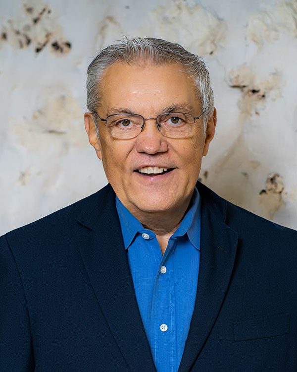 An older man with gray hair and glasses wearing a blue shirt and dark blazer, standing in front of a light-colored, textured background.